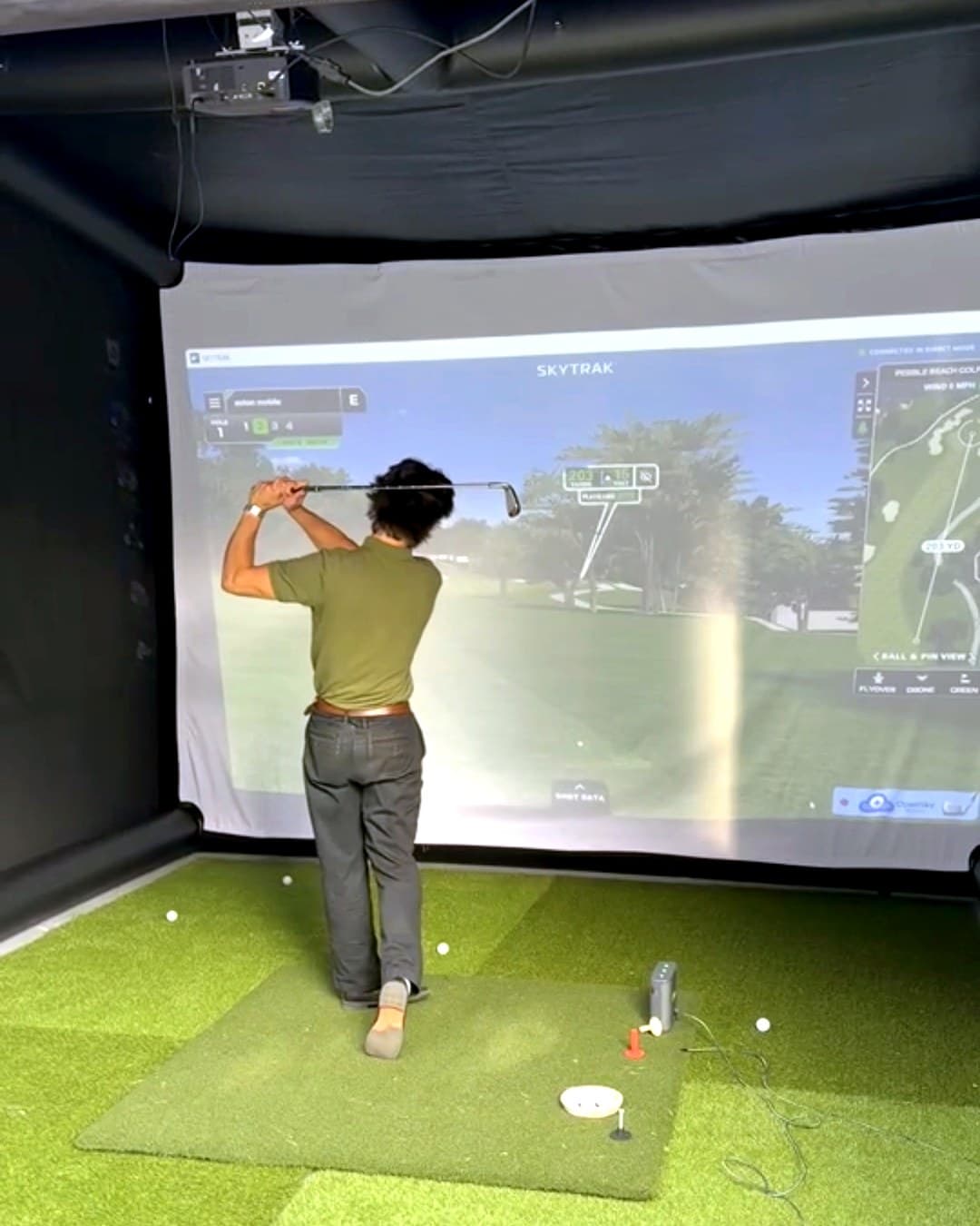 A player mid-swing inside the enclosure, hitting into a full-width projection of a golf hole with shot tracking on screen.